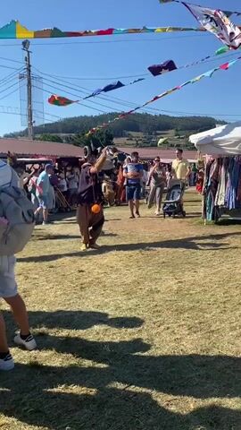 This is a medium shot video taken outdoors on a sunny day. The foreground shows the legs and lower torso of a person wearing shorts and sneakers, facing away from the camera and toward a crowd. In the middle ground, a performer dressed in a medieval-style costume, including a dark tunic, baggy brown pants, and a hat, is juggling orange balls. They are also blowing into a horn. Behind the performer, a crowd of people are milling around a marketplace with various stalls. To the right, a white tent is set up with clothing hanging on display. In the background, there are trees and a hill under a clear blue sky. Colorful triangular flags are strung across the top of the frame.