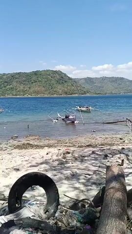 The video shows a beach with debris scattered on the sand. In the foreground, there is a discarded tire and a pile of driftwood and trash. The background features a body of water with several boats. Beyond the water, there are lush green hills under a clear blue sky. The scene appears to be a coastal area that has been affected by pollution.