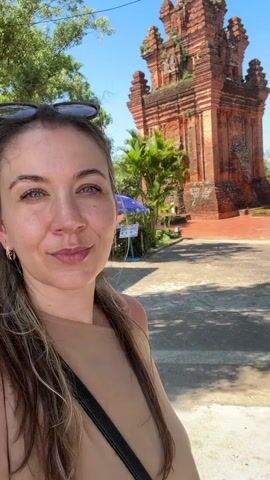 A woman takes a selfie in front of a brick tower. She is wearing a beige top and sunglasses are on top of her head. The tower is tall and looks ancient, with multiple levels. There are trees and a blue umbrella visible behind her. The sky is clear and blue.