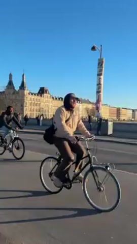 The video shows a bright, sunny day in what appears to be a European city. A man rides a bicycle across a bridge, followed by a group of four people on bicycles. The bridge has a concrete barrier on the left side where pedestrians are walking. In the background, there's a large, ornate building with turrets and a tall street light or post with images on it. The sky is clear blue.