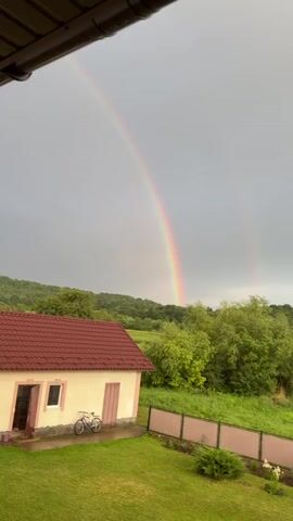 A video captures a rural landscape with a double rainbow arching across a gray sky. In the foreground, a portion of a dark overhang is visible at the top. Below, a small, light-colored house with a red roof sits on the left. A bicycle is parked in front of it. A green lawn stretches out, bordered by a wooden fence. Beyond the house and fence, a patch of green trees and rolling hills covered in foliage form the background. To the right of the house, another brick house is partially visible. The overall scene suggests a post-rain environment.