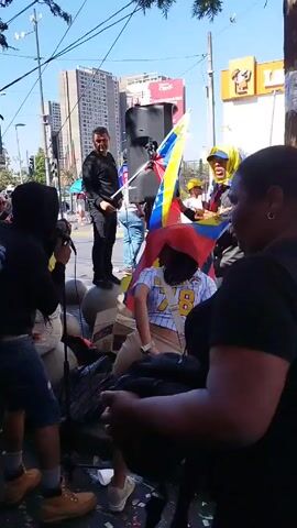 A group of people, some with Venezuelan flags, are gathered in what appears to be a public area. Several individuals are holding and waving the flags.  A man is standing atop a concrete structure behind a speaker. A few people are wearing baseball caps. One person is drinking from a can.  There are buildings in the background. The day is sunny.