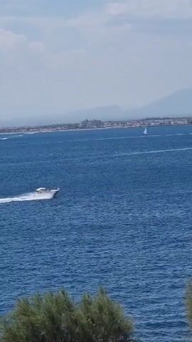 A wide shot of a blue ocean with a distant coastline. A white motorboat speeds across the water in the foreground, leaving a white wake. In the background, a small white sailboat is visible, as is a distant town with buildings and a tall structure, possibly a lighthouse. Mountains are visible in the far distance. The sky is partly cloudy. Green foliage is visible in the bottom foreground.