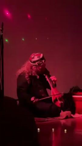 A person is seated on a rug on the floor, playing a bowed string instrument that looks similar to a violin. The person is wearing a hat and dark clothing, and they have long, curly hair. The lighting in the room is red, and there are some green light effects on the wall behind the person. A microphone stand is visible to the left of the person, and a small, red box is behind them. The foreground shows parts of silhouetted viewers.