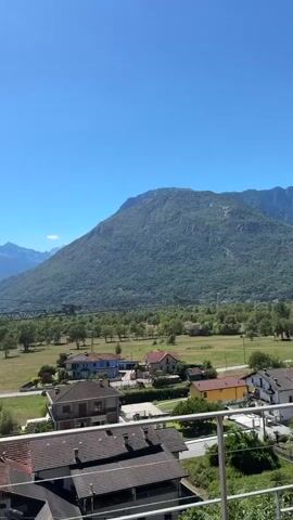 This is a vertical video showcasing a scenic mountainous landscape under a clear blue sky. In the foreground, there is a balcony with a wooden deck and a metal railing. Beyond the balcony, a cluster of houses with red-tiled roofs can be seen. In the middle ground, a wide, green valley stretches out, dotted with trees and some more houses. A prominent crane stands tall in the middle distance, with its boom extended across the valley. Further in the distance, a series of majestic mountains rise, their peaks sharp against the sky. The overall impression is one of a peaceful, rural setting with a backdrop of impressive natural beauty.