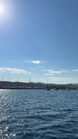 The video captures a scenic view of a sunny bay. The foreground is filled with sparkling blue water, reflecting sunlight, creating a shimmering effect. In the middle ground, numerous sailboats are docked at a marina, their tall masts silhouetted against the bright sky. A few clouds are visible in the distance, adding texture to the clear blue sky. A sailboat with a white mast is sailing on the water. The background features rolling hills covered in lush greenery. The scene is calm and peaceful, suggesting a beautiful day at sea.