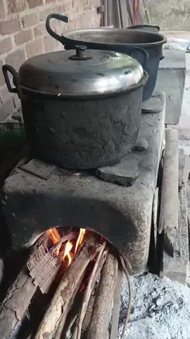 The video shows a close-up shot of a traditional clay stove with a fire burning inside. Two large pots are placed on top of the stove, one in the foreground and another slightly behind it. The pots appear to be made of metal and are blackened with soot. Logs of wood are visible burning inside the stove, with flames reaching upwards. The background shows a brick wall and some wooden structures. The scene suggests a rural or outdoor setting where traditional cooking methods are being used.
