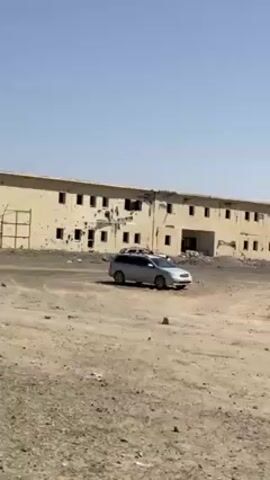 The video shows a two-story building with extensive damage, including holes and cracks in the exterior walls. Several vehicles are parked in front of the building, one of which appears to be heavily damaged. The surrounding area is a dusty, barren landscape under a clear, bright sky. No people are visible in the video. The focus appears to be on documenting the extent of the damage to the building and vehicles.