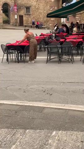 This media file depicts an outdoor scene in a European town square, likely San Quirico d'Orcia, Italy, given the context. The primary subject is a woman wearing a leopard-print jumpsuit and white sneakers, standing at a table with a red tablecloth. She appears to be arranging the tablecloth or setting the table.

The setting is a cobblestone plaza with several tables and chairs, most of which are covered in red tablecloths, suggesting an outdoor dining area for a restaurant or cafe. In the background, there is a stone building with an arched entryway. Two flags, one red and white checkered and another red with white crosses, are visible near the archway. There are other people in the background, some seated at tables under a green awning, and a few walking in the distance.

The activity is the preparation of an outdoor dining space, possibly for lunch or dinner service, with the woman meticulously adjusting the table setting. The time of day appears to be daytime, with natural light illuminating the scene. The weather seems pleasant, suitable for outdoor activities. The overall atmosphere appears calm and perhaps leisurely, typical of a piazza setting in Italy. There is no visible text in the frame.