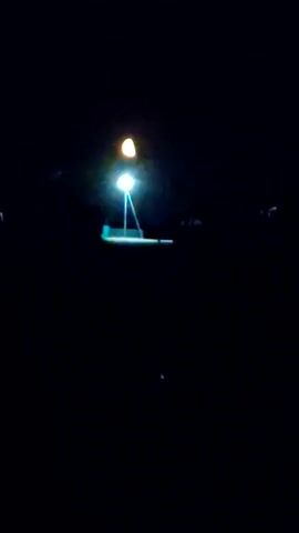 The video shows a dark, outdoor scene at night. The primary focus is a tall, illuminated structure, possibly a light pole, with two bright lights at its apex. One light is a strong white beam, and above it, a softer, yellowish light is visible, suggesting a celestial body like the moon or a distant light source. The pole itself appears to be made of metal with visible supports. Below the lights, a horizontal, illuminated object, possibly a platform or a surface, is partially visible, glowing with a bluish-white light. The background is almost entirely black, indicating a night sky with minimal ambient light. There are faint hints of darker shapes in the periphery, which could be trees or structures, but they are indistinct. The overall impression is of a solitary illuminated object in a dark, open space.