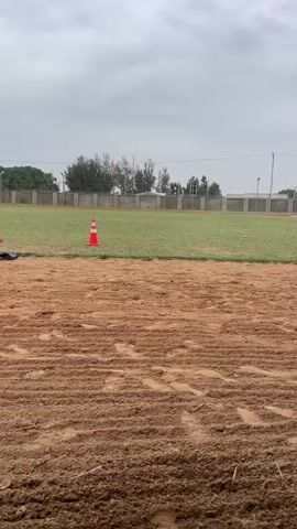 The video shows a man riding a brown horse across a dirt track. The man is wearing a white shirt, black pants, a black cap, and black riding boots. The horse is wearing a saddle and bridle. The track is surrounded by a green field, which is lined with a gray fence. In the background, there are trees and buildings. There are also orange cones scattered around the track. The sky is cloudy and gray.
