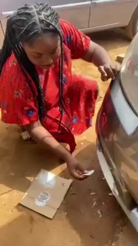 A woman with long black braids is shown crouched down outdoors, applying a white paste to the front bumper of a silver car. She is wearing a red floral dress and striped sandals. A piece of cardboard with blobs of the white paste on it is lying on the ground next to her. The ground is a reddish-brown color. Another silver car is partially visible in the background. The woman is focused on her task, carefully smoothing the paste onto the damaged area of the bumper.