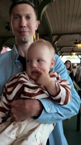 A man holds a baby in his arms. The man is wearing a blue collared shirt. The baby is wearing a striped long-sleeved shirt. The baby's hand is near its mouth. The background appears to be indoors, with wooden ceiling beams visible.