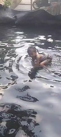 The video shows a person swimming in dark, murky water. The person is partially submerged, and it is difficult to make out their features clearly. The water appears to be stagnant, with ripples and disturbances on the surface. In the background, there is a dark, rocky bank. The lighting is dim, suggesting that the video was taken in low light conditions. There is also a glimpse of what appears to be a motorcycle in the upper right corner of the frame, on the bank.