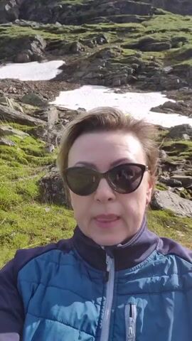 A woman wearing sunglasses and a blue jacket is taking a selfie in a mountainous, grassy area. Snow patches and rocky terrain are visible behind her. The sky is cloudy.