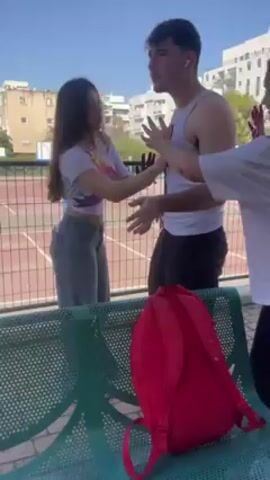 A young man in a tank top is being playfully attacked with colored powder by two young women on a sunny day near a fenced-in tennis court in an urban area. A red backpack and a tube of something are on a green metal bench. All three individuals are smiling and seem to be enjoying themselves. The man's clothes and skin, as well as the women's clothes, are stained with the colored powder.