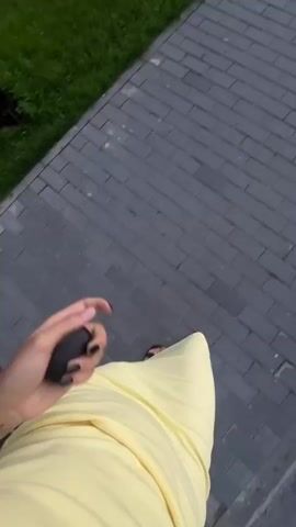 The video begins with a close-up of a person's hand holding a dark object, possibly a phone, with black nail polish. They are wearing a yellow dress and sandals and are walking on a paved walkway next to a grassy area. The camera then pans to show a blurred view of a parking lot with a red car. The scene transitions to a wider shot of a street lined with trees and a fence, with a person walking in the distance on the right side of the frame. The sky is visible with some clouds, and there are buildings in the background.