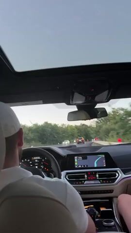 This video shows the interior of a car from the perspective of a passenger in the back seat, looking forward. A person wearing a white cap is driving, with their arm resting on the steering wheel. The car's dashboard displays include a speedometer reading 73, a navigation screen showing a route, and ambient interior lighting. Outside the car, a road is visible with other vehicles ahead, and the sky is overcast. Trees and vegetation line the sides of the road, indicating an outdoor setting. The video appears to be a short clip of a car journey.