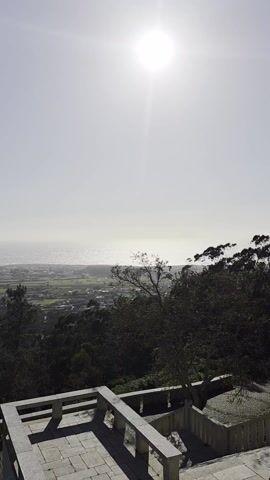The camera is held stationary, capturing a daytime, elevated, wide shot of a landscape. In the foreground, a gray stone patio area with simple white railings is visible. Below the patio, a dense collection of trees forms a green canopy, obscuring much of the land below. Beyond the trees, a patchwork of fields and small buildings stretches towards the coast. In the distance, the ocean meets the horizon under a bright, hazy sky with the sun positioned high and emitting a strong glare. There are no people visible.