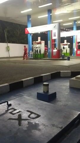 The video shows a gas station in Darungan, Indonesia. The scene is primarily focused on the gas pumps and the canopy above them. A person wearing a red uniform, likely a gas station attendant, is visible near one of the pumps. The time of day appears to be evening or night, as indicated by the artificial lighting illuminating the station. The weather seems clear.

Notable objects include the gas pumps themselves, with branding visible. The canopy has prominent red and white trim, and a white ceiling with multiple fluorescent lights. The ground is paved, with black and white striped markings along a curb. To the right of the frame, the ground is concrete and has graffiti written on it. The graffiti appears to be in black marker and includes the letters "PT" and what looks like a stylized "M" or "W". There is also a blue, T-shaped object on the concrete.

In the background, beyond the gas station, trees are visible, suggesting an outdoor setting. The overall atmosphere is functional and quiet, characteristic of a gas station at night. There are no strong emotional cues conveyed. Text visible includes "PERTAMINA DEX" and "PERTAMAS" on the gas pumps, indicating the type of fuel offered. There is also a "1" on a red rectangular sign near a pump.