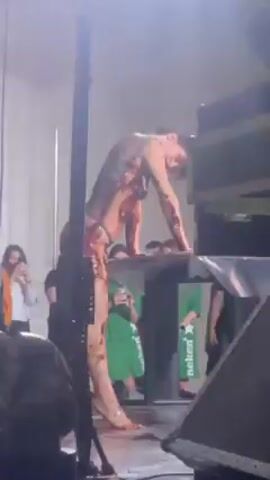 The video shows a shirtless person with dark hair wearing a short, patterned skirt and no shoes, dancing on a platform. The person dances close to a stack of black equipment, with a green Heineken box visible at the bottom. The person bends over at the waist, facing away from the camera, and moves their hips rhythmically. Several blurred figures stand in the background, seemingly watching the performance. The setting appears to be an indoor venue, with tall white curtains visible on the left side. The lighting is bright, with a chandelier visible in the background.