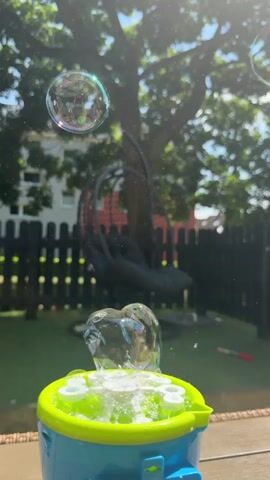 A bubble machine is in the foreground, emitting bubbles into the air. The bubble machine is blue with a lime green top and is on a surface that appears to be a table. Bubbles are rising from the machine and floating around it. The background shows a backyard with a green lawn, a black wooden fence, and a large tree with green leaves. There is also a hanging chair visible in the backyard. The sun is shining, casting rays of light through the bubbles.