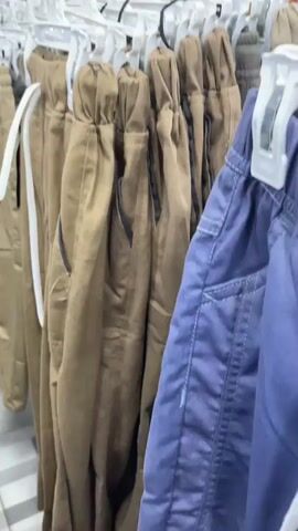 This is a video taken inside a clothing store. The camera pans across rows of pants hanging on display racks. The pants are mostly a tan or brown color, with some dark blue jeans visible on the right side of the frame. The pants appear to be children's clothing, as evidenced by the small size and decorative embroidery on some of the jeans, such as flowers and a pineapple. The store seems to be well-stocked, with many items on display. In the background, a woman wearing a hijab and a yellow shirt is browsing the clothing racks. The floor is tiled with a gray and white striped pattern. The lighting in the store is bright, suggesting overhead fluorescent lights. The video captures a typical retail environment, with clothing neatly organized and displayed for sale.