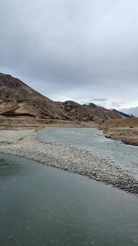 A wide, shallow river flows through a valley with rolling hills on either side. The hills are covered in dry grass and scrub, with some patches of snow on the higher peaks in the distance. The sky is overcast with dark clouds. The water is a murky green color, and there are rocks and gravel visible in the riverbed. The video pans slowly from left to right, showing the landscape in a wide shot.