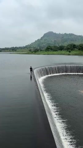 A person walks along the edge of a circular weir, a man-made structure over which water cascades in a semi-circular curtain. The person is silhouetted against the calm water of a reservoir, with a green, tree-covered hill forming the backdrop under a cloudy sky. The water on the weir is turbulent, contrasting with the stillness of the reservoir. The person is walking away from the camera towards the weir.