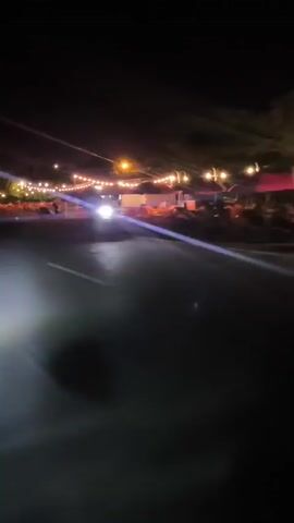 A nighttime video shows a roadside view of a restaurant with outdoor seating illuminated by string lights. The camera is moving, giving a sense of passing by the establishment. Numerous empty tables and chairs are visible, arranged in rows under a covered area. A few motorcycles are parked near the restaurant. The road in the foreground is dark, with the headlights of a vehicle creating bright streaks of light. The sky is black, indicating it is late at night. There are no people clearly visible in the frame, suggesting the establishment may be closed or not busy.
