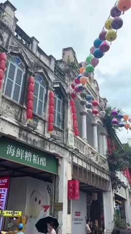 This is a vertical video of a street lined with old buildings adorned with strings of colorful lanterns. The buildings appear to be made of concrete or stucco and have architectural details such as columns and arched windows. Many of the buildings have signs in Chinese characters. Red lanterns are also visible, hanging vertically along the facades of some buildings. The street is bustling with people walking and some are sitting at outdoor tables. The sky is overcast. The camera pans from left to right, showing more of the street and the buildings.