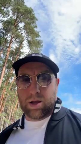 A man wearing a black baseball cap with an Under Armour logo, sunglasses, and a black jacket over a white t-shirt is walking outdoors. He is facing the camera and appears to be speaking, with his mouth open. The setting is a paved pathway lined with tall, slender pine trees. To the left of the path, there is a neatly manicured lawn. The sky is blue with scattered white clouds, suggesting it is daytime. The lighting indicates it is likely a sunny day. The pathway is made of light-colored pavers. In the background, beyond the trees, a large expanse of green lawn stretches out, and there are some white decorative elements that could be sculptures or flower beds. The overall impression is of a well-maintained, possibly park-like or estate-like environment. The location context provided is Мядзельскі сельскі Савет, Belarus, which places this scene in a rural or semi-rural area of Belarus.