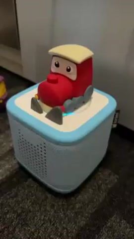 The video shows a red, cartoon train figure sitting on top of a light blue Toniebox speaker. The speaker is placed on a patterned rug.  The train figure has a cream-colored roof, large eyes, and grey wheels and shoes. The Toniebox begins to glow with an orange light, and a voice speaking in German is heard. Another cartoon figure is partially visible to the left of the speaker. A mirrored surface reflects light in the background.
