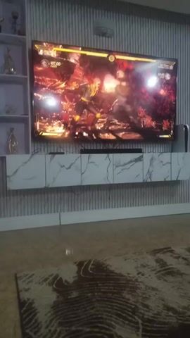The video shows a large flat-screen television displaying a scene from the Mortal Kombat game. Two characters are engaged in combat within the game. The television is mounted on a wall above a white cabinet with a marble pattern. A speaker bar is placed on the cabinet beneath the television. The wall has a textured, vertical line design. The floor appears to be a glossy, light-colored surface. A bookshelf is partially visible to the left of the television.  The characters in the game are fighting in a fiery, cave-like environment. The health bars of the characters and the game's interface are visible at the top of the screen. The lighting in the room is bright.