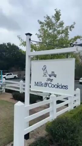 A video shows an establishment called "Tiny's Milk & Cookies."  The building is a small white structure with a dark roof, situated behind a white picket fence.  A sign reading "Tiny's Milk & Cookies" hangs from a post near the fence. A few tables and chairs sit on a patio near the building. Several people are standing in line at the window of the building, presumably waiting to order. Behind the building, vehicles are parked. The sky is overcast.