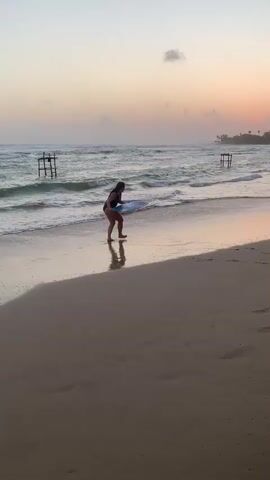 A lone person in a dark swimsuit is on a sandy beach at dawn. They are bent over, facing the ocean, and appear to be interacting with a round, flat object on the wet sand near the water's edge. Gentle waves are breaking on the shore. In the background, two wooden structures rise from the water. The sky is a gradient of pale pink and orange, with a few wispy clouds. The overall impression is one of tranquility and solitude at the beginning of a new day.