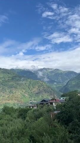 The video shows a sweeping view of a valley in the mountains. The sky is a vibrant blue with fluffy white clouds. Lush green mountains rise in the background, their slopes dotted with trees and some small buildings. In the foreground, a dense forest of green trees covers the hillside. A few houses with dark roofs are visible nestled amongst the trees, hinting at a small settlement within the valley. The overall impression is one of natural beauty and tranquility.