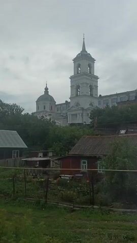 The video shows a view of a village or town with a prominent white bell tower and a dome structure in the background. In the foreground, there are several residential buildings, including houses with red and blue roofs, and fences. Green vegetation, including trees and grass, is abundant throughout the scene. A white SUV is parked on a dirt road. The sky is overcast. The camera appears to be moving slightly, possibly from a vehicle.