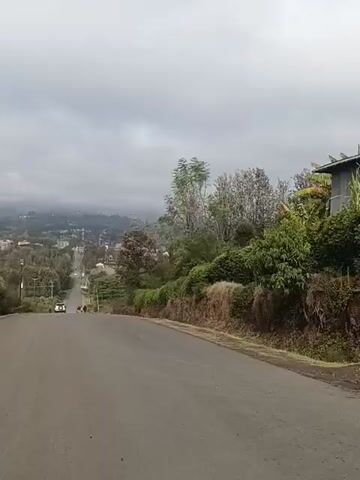 The camera pans down a paved road in what appears to be a mountainous region. The day is overcast with a thick cloud cover. The road descends into a valley with a visible town in the distance. Green vegetation lines the side of the road, and a few people are walking down the street. There are also several vehicles in the distance on the road.