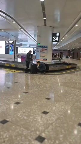 The video captures the interior of a modern airport baggage claim area, likely in Langfang, China. At the start, two men are visible near a baggage carousel; one stands while the other crouches to manage luggage. The carousel has yellow marked areas on the floor in front of it. In the background, large digital displays show Chinese text, with one display featuring phrases like "华为云码道" (Huawei Cloud Code Way) and "要懂AI编程" (Need to understand AI programming). Another display shows a scenic landscape image. The number "34" is clearly displayed on overhead signs, indicating the carousel number. As the camera pans to the right, a long, neatly arranged row of silver luggage trolleys fills the frame. Further in the background, an "出口 Exit" sign, written in both Chinese and English, directs passengers. The airport features a high, curved ceiling with recessed lighting and a polished tiled floor with a grey and black pattern. The overall environment is well-lit and appears to be clean and organized.
