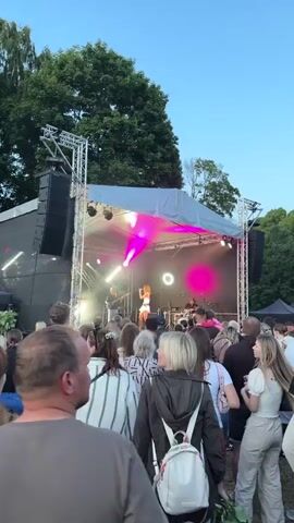 The video shows a live outdoor music performance. A female singer is on stage, wearing a white tank top and a short white skirt, with her hair down. She is singing and dancing. Behind her, a band is performing, with a guitarist to her left and a drummer to her right. The stage is set up with professional lighting, including bright pink and white spotlights that illuminate the performers and create beams of light. The stage is covered by a white canopy.

In front of the stage, a crowd of people is gathered, watching the performance. They are standing and some are holding up their phones to record or take pictures. The audience appears diverse in age and gender. The lighting and the clear sky suggest it is evening, possibly during the summer. The presence of trees in the background indicates an outdoor park or open field setting. The atmosphere appears to be festive and energetic, with the audience engaged by the music. The location is identified as Panevėžys, Lithuania.