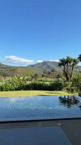 The camera is stationary and shows a view from a pool in a lush, tropical setting. The pool's water reflects the blue sky and hints of clouds. Beyond the pool is a green lawn, followed by various trees and shrubs. In the distance are hills or mountains under the clear sky. The overall impression is that the setting is serene and natural. The edge of a lounge chair is visible at the bottom of the frame. Towards the end of the clip, orange-colored planters are revealed.