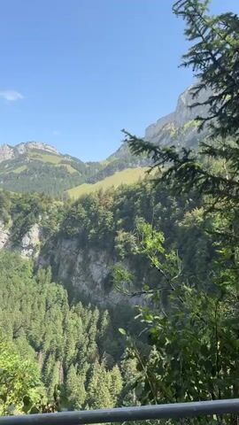 The video shows a mountainous landscape with lush green trees and rocky cliffs. The sky is a clear blue with a few wispy clouds. The camera pans from a wide shot of the mountains to a closer view of a hiking trail that winds along a cliff face. Several people are seen walking on the trail, enjoying the scenery. In the foreground, the leaves of trees obscure some of the view. The overall impression is of a beautiful and serene natural environment.