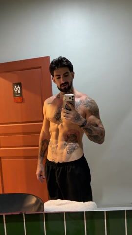 A muscular man with a beard is standing in a bathroom taking a selfie in the mirror with his smartphone. He is shirtless, wearing only black shorts, and has several tattoos on his chest, arms, and abdomen. He is smiling slightly at the phone. Behind him is an orange door with a restroom sign, a white wall, and green tiled trim along the bottom of the mirror. There's a dark object, possibly a chair, to his left.