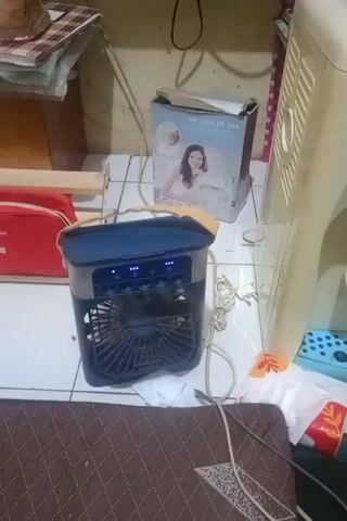 A personal air cooler fan is placed on the floor. The fan is dark blue and gray with a clear handle on top. It has a mesh front with a visible fan blade. The fan is plugged in with a white cord. Next to the fan, there is a box with an image of a woman on it, labeled "AIR COOLER FAN". There are also some scattered items, including what appears to be a red bag, some papers on a shelf, and a portion of a water dispenser. The floor is tiled. The lighting is dim.