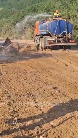 The video depicts a construction site, likely a road being built or maintained. The foreground shows a muddy, dirt road with tire tracks and scattered rocks. A person in a yellow safety vest and a hard hat is partially visible on the left, holding a wooden pole, suggesting they are involved in the construction work.

In the background, a large orange and blue water truck is actively spraying water onto the road surface. A person in a safety vest and hard hat is standing on top of the truck, operating the water spray mechanism. The purpose of watering the road is likely to suppress dust during construction, compact the soil, or prepare it for paving.

The setting appears to be a rural or undeveloped area, with a hillside covered in green vegetation visible behind the truck. The lighting suggests it is daytime, with sunlight casting shadows on the ground. The overall scene conveys an active construction environment.

There is some visible text on the back of the truck, including a license plate that appears to read "HUY 303" and other illegible markings. The presence of safety gear on the workers indicates a focus on occupational safety.