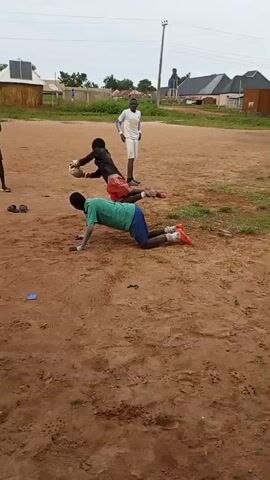 A group of young men are practicing soccer drills on a dirt field. In the foreground, one young man in a green shirt and blue shorts is on his hands and knees with a soccer ball between his legs. Another young man in goalkeeper gloves and pads is lying on his back with his legs extended. A third young man stands watching in the background, wearing a white shirt and shorts. Another young man in a black shirt and shorts stands to the left, also observing. The field is dry and dusty, with some patchy grass. In the background, there are some simple structures, including what appears to be a small building with solar panels on the roof, and other houses. The sky is overcast. The action shows the young man on his hands and knees attempting to control the ball with his feet while the other person on the ground also interacts with the ball.