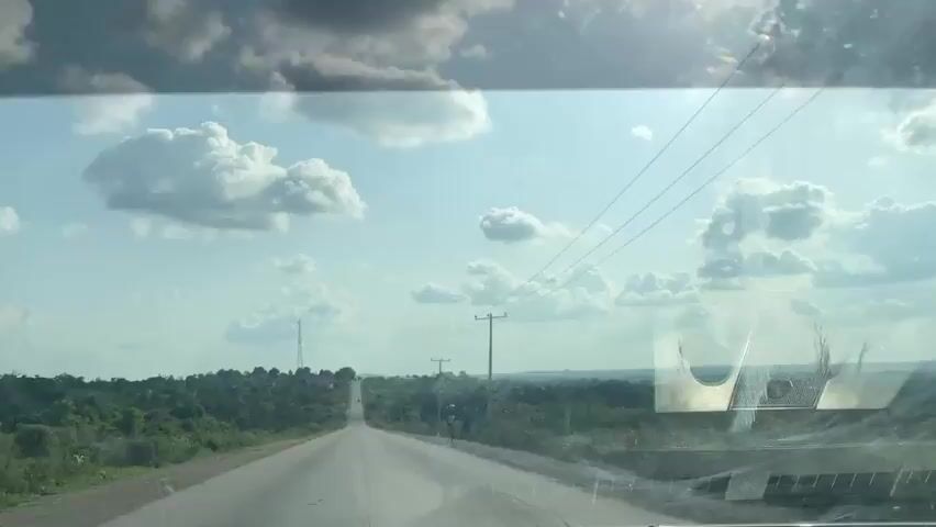 This media file captures a view from inside a vehicle, looking out onto a long, straight, paved road in a rural setting. The road stretches towards the horizon, lined with lush green vegetation on either side. In the distance, a town or village is visible, with buildings clustered together. A tall communications tower stands out against the sky.

The sky is a bright blue, dotted with fluffy white cumulus clouds, suggesting it is daytime, likely mid-morning or afternoon, with fair weather. Sunlight streams down, creating a bright atmosphere. Power lines stretch across the scene, supported by wooden utility poles, indicating infrastructure in the area.

Two individuals are seen walking on the side of the road in the middle distance, adding a sense of scale and human presence to the landscape. The overall mood is one of travel and exploration through a scenic, undeveloped region. The reflection on the windshield includes a metallic object, possibly a rearview mirror or part of the car's interior, with a circular imprint visible. There is no visible text in the media file. The location context provided is Oduape, Nigeria.