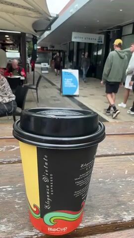 A coffee cup sits on a wooden table in the foreground. In the background, a group of people walk down a covered walkway lined with shops. One shop is labeled "JETTY STRATA." A blue sign with white text reads "A good day everything." A young boy in a blue shirt is visible on the right side of the frame, facing away from the camera and reaching out towards the right.