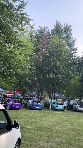 This media file captures an outdoor car meet event held in a park-like setting, likely in Târgu Ocna, Romania. The scene is filled with various customized cars, many with their hoods or trunks open, suggesting a display or competition. Several tents are set up, some with visible branding, indicating sponsors or vendors at the event.

People are seen scattered throughout the area, some gathered around the cars, others sitting and observing, and a few walking. The atmosphere appears relaxed and social. The time of day seems to be late afternoon or early evening, with the sun casting long shadows and a soft light filtering through the abundant trees that surround the gathering. The weather appears to be pleasant, with a clear or partly cloudy sky.

Several cars are prominent in the foreground and mid-ground, including a bright purple hatchback, a blue hatchback, and a teal sedan. Towards the back, a silver car and a black SUV are also visible.

Text visible in the media includes branding on the tents. One tent prominently displays "ALPHARD" in large white letters, with additional text below that is partially obscured. Another tent has "Reis..." visible. A banner within one of the tents reads "EBEDRAW" along with several logos. The general theme of the event seems to be car modification and enthusiast gathering. The overall impression is a community event celebrating automotive culture in a natural, green environment.